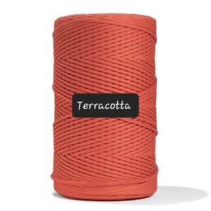 May include: A cylindrical spool of terracotta-colored macrame cord. The cord is tightly wound, showing a textured surface. A black rectangle with the word "Terracotta" in white text is at the bottom of the spool.
