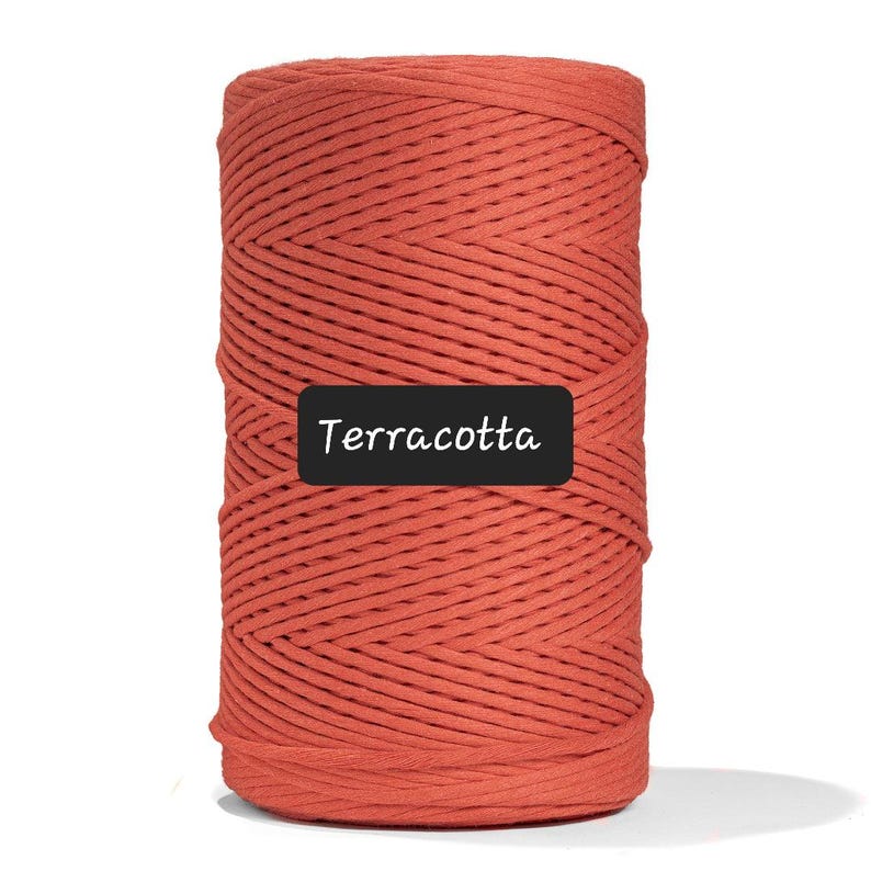 May include: A cylindrical spool of terracotta-colored macrame cord. The cord is tightly wound, showing a textured surface. A black rectangle with the word "Terracotta" in white text is at the bottom of the spool.