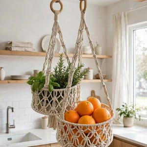 May include: A two-tiered macrame hanging basket made of off-white cotton cord. The top basket holds green herbs, while the bottom basket is filled with bright orange citrus fruits. Each basket features a knotted lattice design and a tassel.