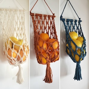 May include: Three macrame hanging fruit baskets suspended from wooden dowels. Each basket features a knotted net design, a bottom tassel, and a hanging cord. The baskets are filled with yellow lemons and orange citrus fruits in cream, rust, and navy blue.