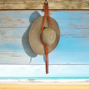 May include: A brown woven sun hat with a brown leather strap hanging from a wooden beam against a blue and white wood background.