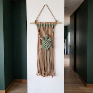 May include: A macrame wall hanging featuring a central sage green turtle design crafted from knotted cord. The piece is suspended from a horizontal wooden dowel by a tan rope hanger, with long, loose tan fringe strands hanging below the turtle.