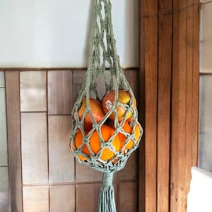 May include: A hanging macrame fruit hammock in a muted green color, filled with oranges and apples. The text "Style Twisted" is displayed in red at the top. The hammock is suspended from a wooden ring.