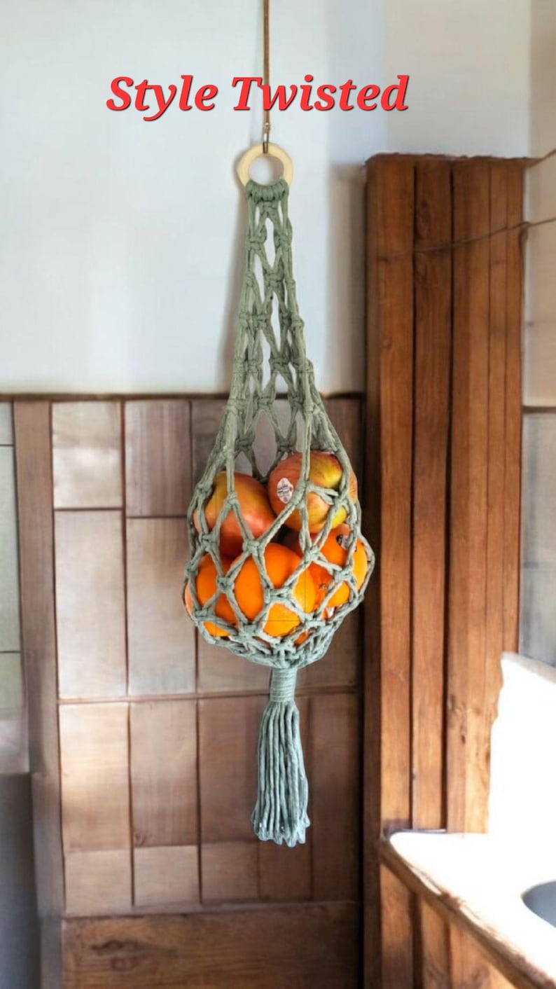 May include: A hanging macrame fruit hammock in a muted green color, filled with oranges and apples. The text "Style Twisted" is displayed in red at the top. The hammock is suspended from a wooden ring.