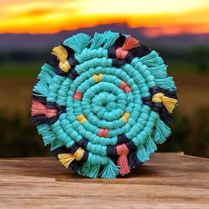 May include: A round, turquoise macrame coaster with a spiral design. The coaster features accents of black, yellow, and coral-colored fringe. The coaster is sitting on a wooden surface with a blurred sunset background.