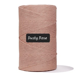 May include: A spool of dusty rose-colored macrame cord. The cord is tightly wound, showing a textured surface. A black label with the words "Dusty Rose" is affixed to the front of the spool. This cord is suitable for various crafting projects.