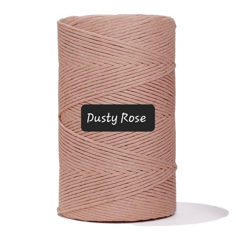 May include: A spool of dusty rose-colored macrame cord. The cord is tightly wound, showing a textured surface. A black label with the words "Dusty Rose" is affixed to the front of the spool. This cord is suitable for various crafting projects.