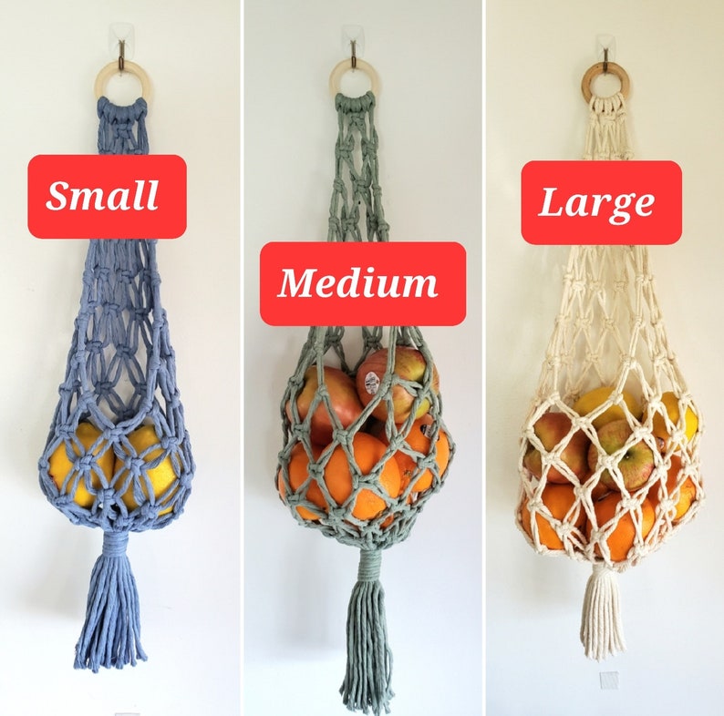 May include: Three macrame fruit hammocks in different sizes: small, medium, and large. The small hammock is blue, the medium is green, and the large is off-white. Each hammock has a wooden ring at the top and a tassel at the bottom. The hammocks are filled with fruit.