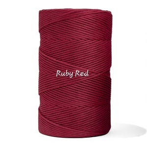 May include: A spool of ruby red macrame cord. The tightly wound cord is a deep, rich red color. The text "Ruby Red" is written in a cursive font across the front of the spool. The cord is suitable for various crafting projects.