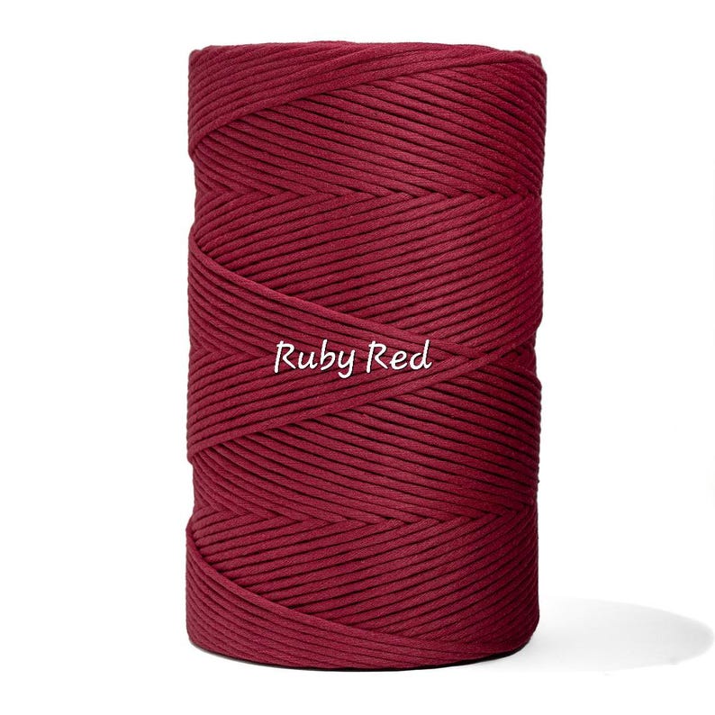 May include: A spool of ruby red macrame cord. The tightly wound cord is a deep, rich red color. The text "Ruby Red" is written in a cursive font across the front of the spool. The cord is suitable for various crafting projects.