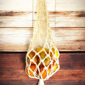 May include: A cream-colored macrame fruit bag filled with oranges and apples, hanging against a wooden background. The bag has a wooden ring at the top for hanging and a tassel at the bottom. The text "Style Twisted" is at the top.