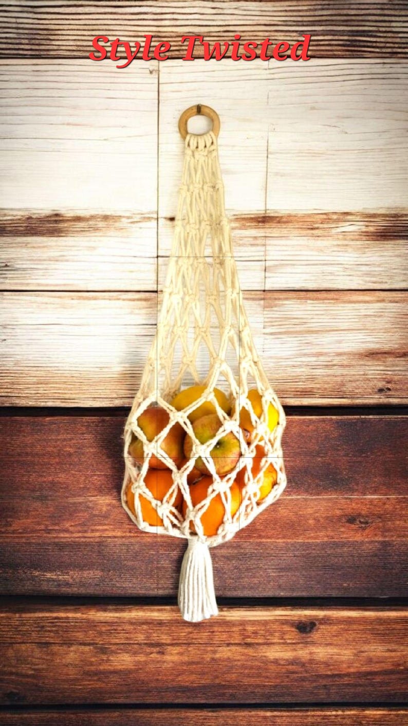 May include: A cream-colored macrame fruit bag filled with oranges and apples, hanging against a wooden background. The bag has a wooden ring at the top for hanging and a tassel at the bottom. The text "Style Twisted" is at the top.