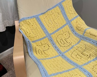 May include: A crocheted blanket with a yellow and blue checkered pattern. The blanket is draped over a light brown armchair.