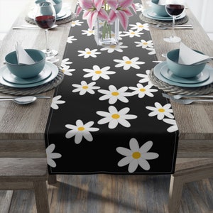 May include: A black table runner with a white daisy pattern. The runner is decorated with white daisies with yellow centers. The runner is on a wooden table with two place settings on each side.