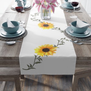 May include: White table runner with a sunflower design. The runner features a vine with green leaves and yellow sunflowers with brown centers. The runner is on a wooden table with two place settings on each side.