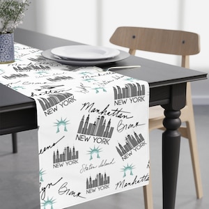May include: White table runner with a black and white repeating pattern of the New York City skyline, the Statue of Liberty, and the words "Manhattan", "Bronx", and "New York".