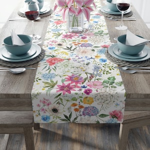 May include: A white table runner with a floral pattern featuring pink, blue, yellow, and green flowers. The runner is on a wooden table with two place settings on each side.