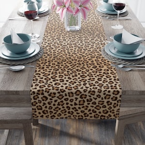 May include: A brown and tan leopard print table runner with a subtle ombre effect. The runner is laid out on a wooden table with two place settings on each side.