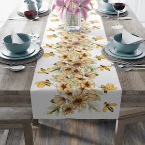 May include: White table runner with a yellow and green floral design featuring sunflowers and bees. The runner is on a wooden table with two place settings on each side.