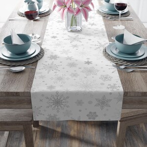 Puede incluir: Un camino de mesa blanco con un patrón repetido de copos de nieve plateados. El camino de mesa está colocado sobre una mesa de madera, con cubiertos, copas de vino y un jarrón con flores rosas. La mesa está puesta para una comida festiva.
