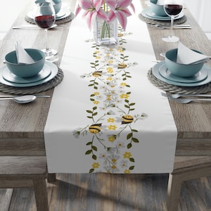 May include: White table runner with a repeating pattern of bees and yellow and white flowers on a green vine. The runner is on a wooden table with a vase of pink flowers in the center.