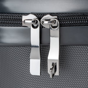 May include: Close-up of a silver zipper on a gray fabric suitcase. The zipper has two pulls and is secured with a small metal loop.
