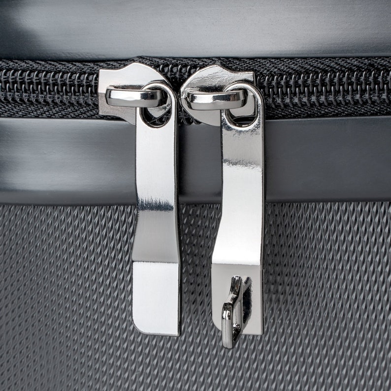 May include: Close-up of a silver zipper on a gray fabric suitcase. The zipper has two pulls and is secured with a small metal loop.