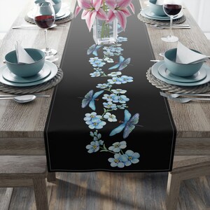 May include: Black table runner with a white border, adorned with a repeating pattern of blue butterflies and forget-me-not flowers. The runner is set on a wooden table, with place settings and a vase of pink lilies.