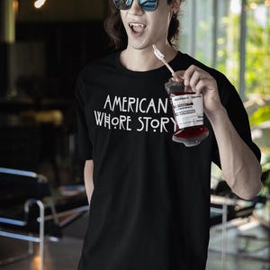 May include: A black short-sleeved t-shirt featuring the white text 'AMERICAN WHORE STORY' printed on the chest. The shirt is worn by a person with dark hair, sunglasses, and visible vampire-style fangs, holding a plastic blood bag with a straw.