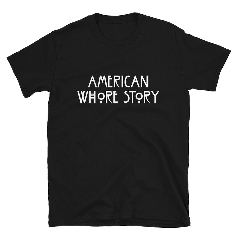 May include: A black short-sleeved t-shirt featuring the white text AMERICAN WHORE STORY printed in a stylized, clean font across the center of the chest. The garment has a crew neckline and a standard fit, showcasing a bold graphic design.