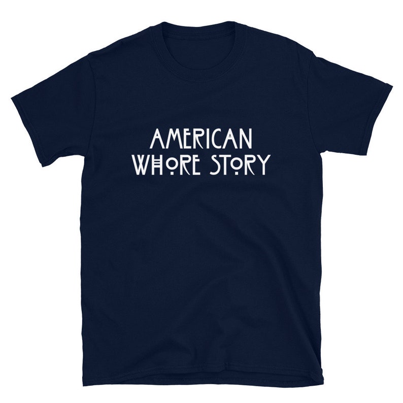 May include: A dark navy blue short-sleeved t-shirt featuring the white text AMERICAN WHORE STORY printed in a stylized, clean font across the center of the chest. The garment has a crew neckline and a standard, straight-cut silhouette.