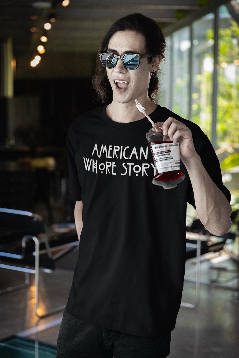 May include: A black short-sleeved t-shirt featuring the white text 'AMERICAN WHORE STORY' printed on the chest. The shirt is worn by a person with dark hair, sunglasses, and visible vampire-style fangs, holding a plastic blood bag with a straw.