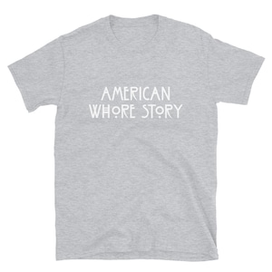 May include: A heather grey short-sleeved t-shirt featuring the white text AMERICAN WHORE STORY printed in a stylized, sans-serif font across the center of the chest. The garment has a crew neckline and a standard, straight-cut silhouette.