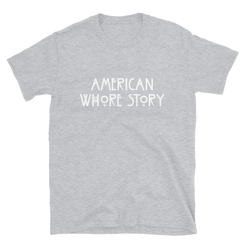 May include: A heather grey short-sleeved t-shirt featuring the white text AMERICAN WHORE STORY printed in a stylized, sans-serif font across the center of the chest. The garment has a crew neckline and a standard, straight-cut silhouette.