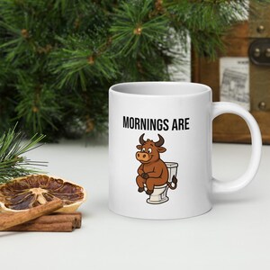 Könnte beinhalten: Weiße Keramik-Tasse mit dem Text "MORNINGS ARE" und einem Cartoon-Bullen auf einer Toilette. Der Bulle ist braun mit schwarzen Hörnern. Die Tasse hat einen weißen Griff.