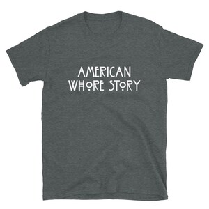 May include: A dark heather grey short-sleeve t-shirt featuring the white text AMERICAN WHORE STORY printed in a stylized, geometric font across the center of the chest. The garment has a crew neckline and a standard, straight-cut silhouette.