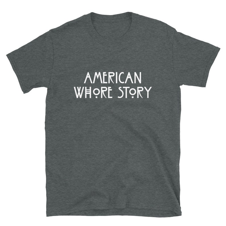 May include: A dark heather grey short-sleeve t-shirt featuring the white text AMERICAN WHORE STORY printed in a stylized, geometric font across the center of the chest. The garment has a crew neckline and a standard, straight-cut silhouette.