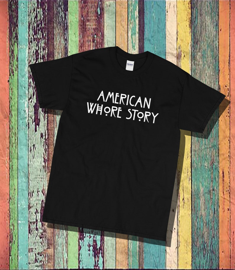 May include: A black short-sleeved t-shirt featuring the white text AMERICAN WHORE STORY printed in a stylized font across the chest. The garment has a standard crew neckline and a visible brand label at the inner collar area.