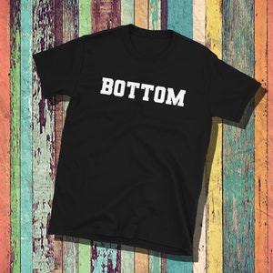 Puede incluir: Camiseta negra con la palabra "BOTTOM" impresa en letras mayúsculas blancas y en negrita. La camiseta es de cuello redondo y parece estar hecha de un material suave. El fondo es una superficie de madera colorida y desgastada.