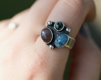 May include: A silver ring with three different colored stones. The stones are a brown agate, a blue agate, and a green agate.