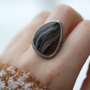 May include: A silver ring with a large, teardrop-shaped stone. The stone features a swirling pattern of gray and brown hues, set within a scalloped silver bezel. The ring is a piece of jewelry.
