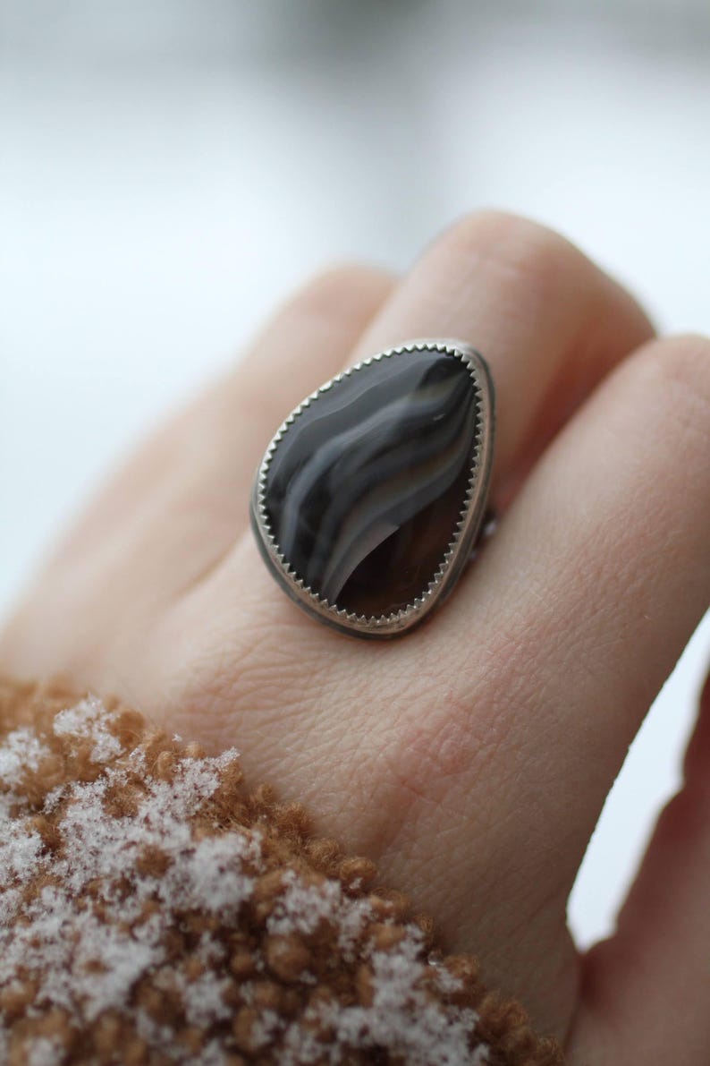 May include: A silver ring with a large, teardrop-shaped stone. The stone features a swirling pattern of gray and brown hues, set within a scalloped silver bezel. The ring is a piece of jewelry.