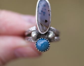 May include: A silver ring with a blue and gray agate stone and a smaller blue stone. The ring is set in a silver band with a textured design.