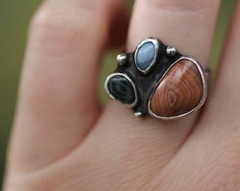 May include: A silver ring with three different colored stones. The stones are a light blue, a dark green, and a brown with white stripes.