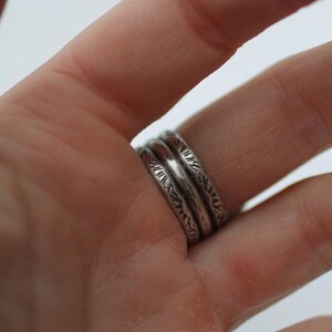 May include: Close-up of three silver rings with textured designs. The rings are stacked on a finger, showcasing the intricate patterns and the metallic sheen of the jewelry. The rings have a rustic, handcrafted appearance.