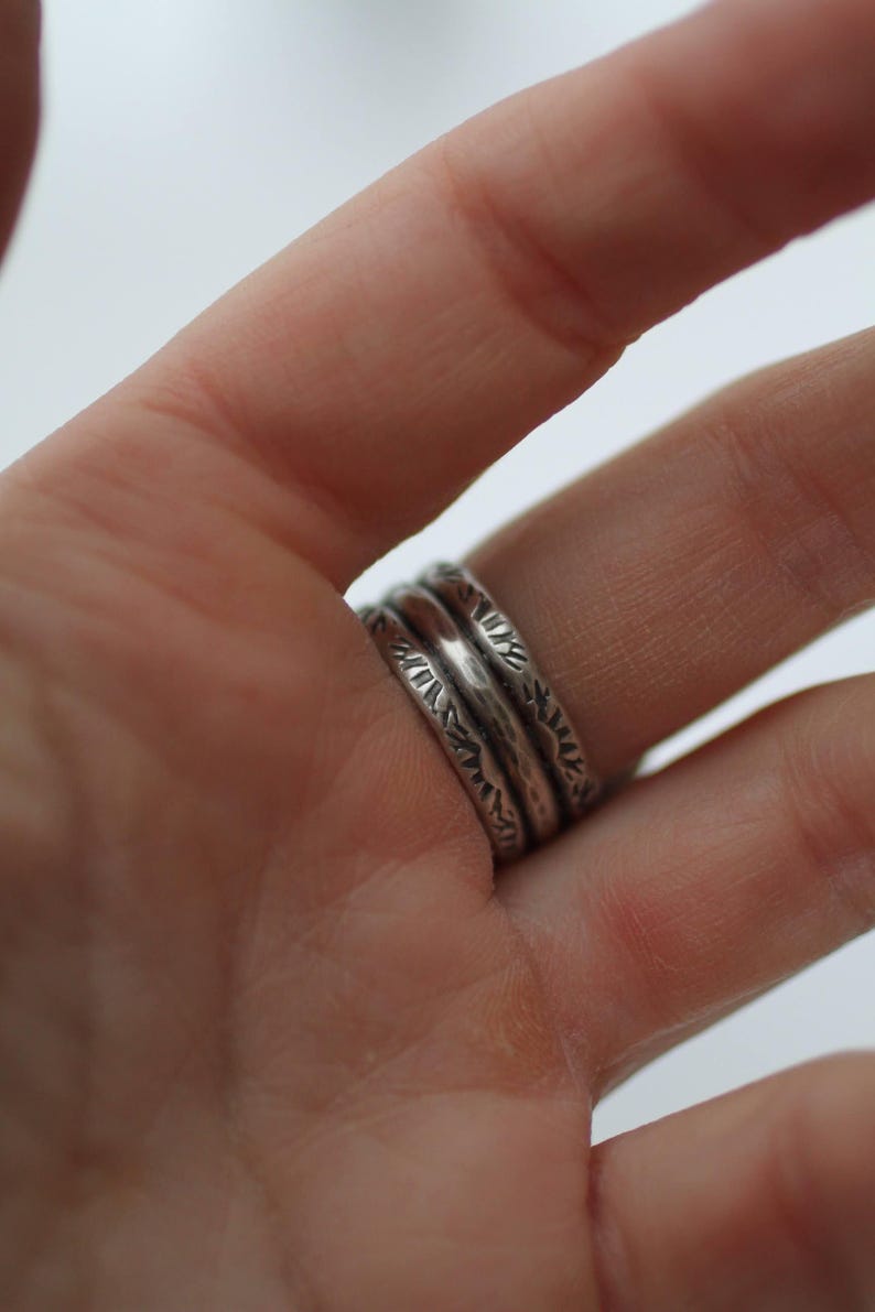 May include: Close-up of three silver rings with textured designs. The rings are stacked on a finger, showcasing the intricate patterns and the metallic sheen of the jewelry. The rings have a rustic, handcrafted appearance.