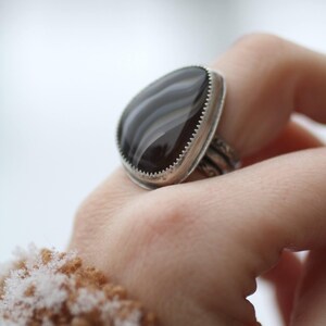 May include: A silver ring with a large, teardrop-shaped, black and gray agate stone. The stone is set in a silver bezel with a serrated edge. The ring's band has a detailed, textured design.