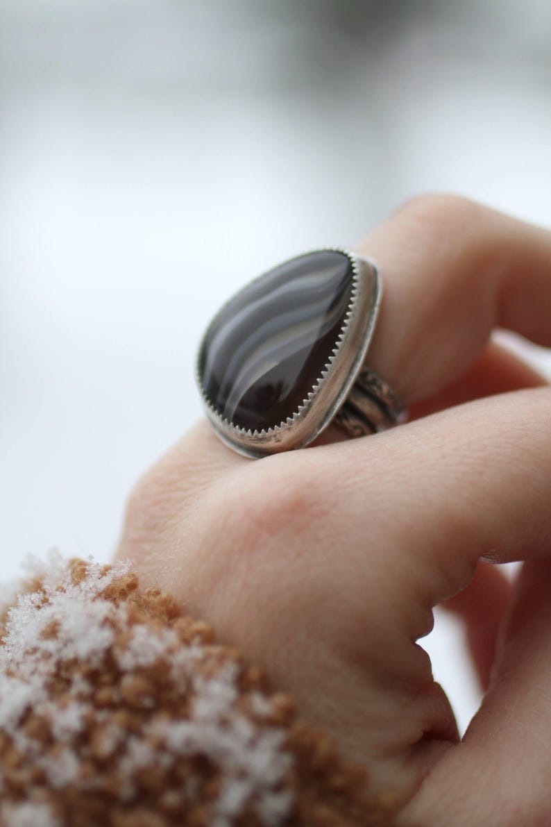 May include: A silver ring with a large, teardrop-shaped, black and gray agate stone. The stone is set in a silver bezel with a serrated edge. The ring's band has a detailed, textured design.