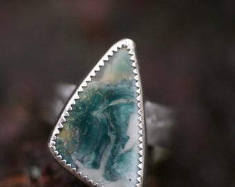 May include: A silver ring featuring a triangular, natural stone with a unique pattern of green, white, and yellow hues. The stone is set within a serrated silver bezel, creating a striking contrast.