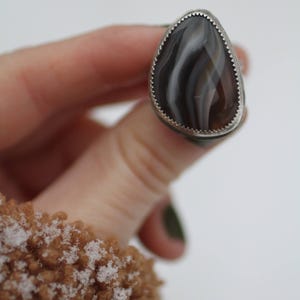 May include: A silver ring with a teardrop-shaped stone. The stone features a pattern of brown, gray, and white bands. The ring has a serrated edge around the stone.
